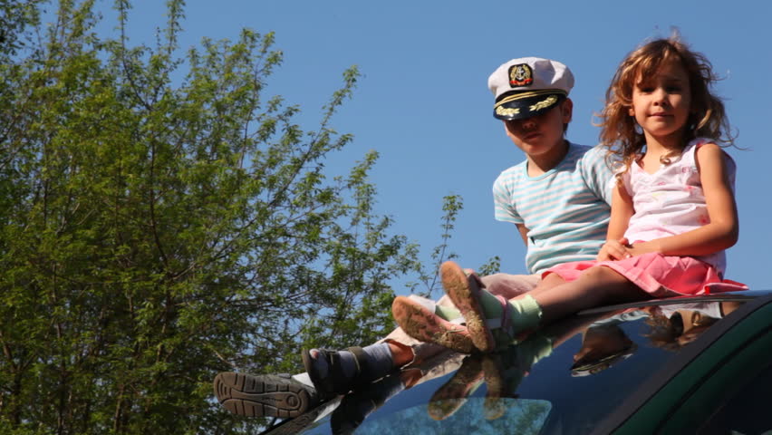 boy in captain's cap and his little sister sit on roof of car near wood against blue sky, she looks at sun covering himself hand