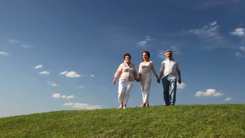 mature couple holding hands with their adult daughter go down grassy hill and leave against sky in summer