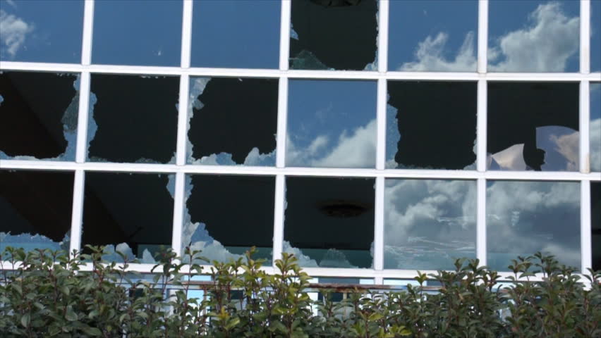 A blue sky with fluffy white clouds moving rapidly across it, reflected in the numerous panes of a large glass wall, many of which are broken.