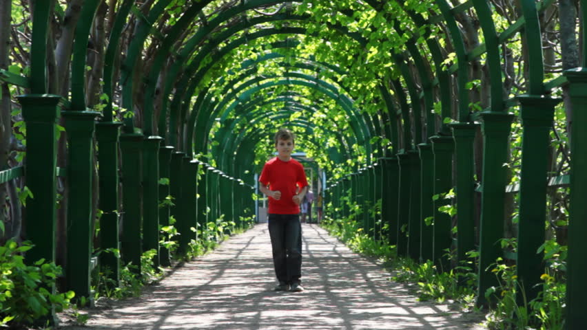 boy in red shirt runs through arched corridor braided green plants by spring day
