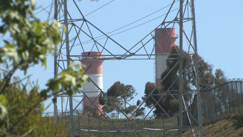 Red & white industrial smoke stacks at an oil refinery. Power lines on a hill near the coast of California. A tree can be seen blowing in the wind in the foreground. Peaceful scene. 