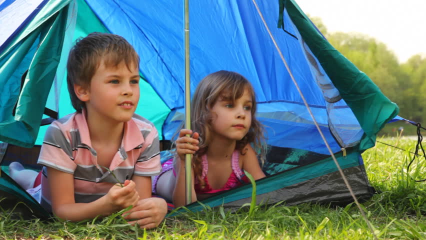 boy with little girl lay peeping out of tent, he something says at forest on summer day