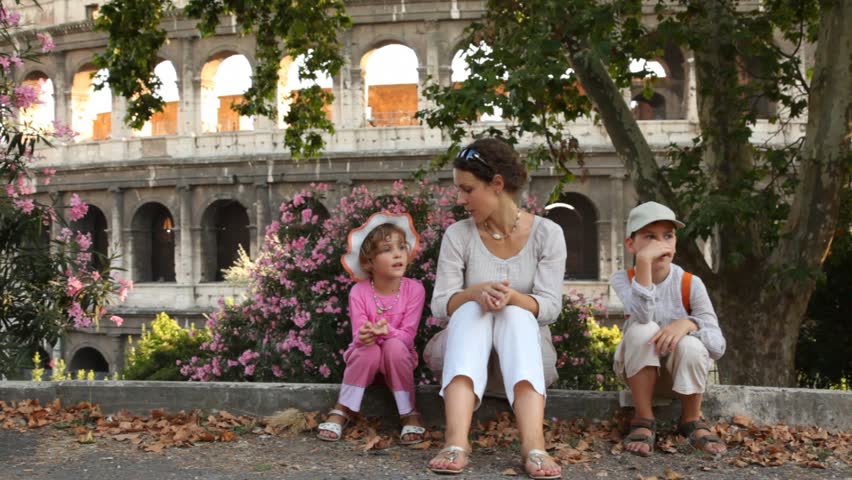 woman tells story to girl and boy sit under shade of tree in front of Colosseum in Rome