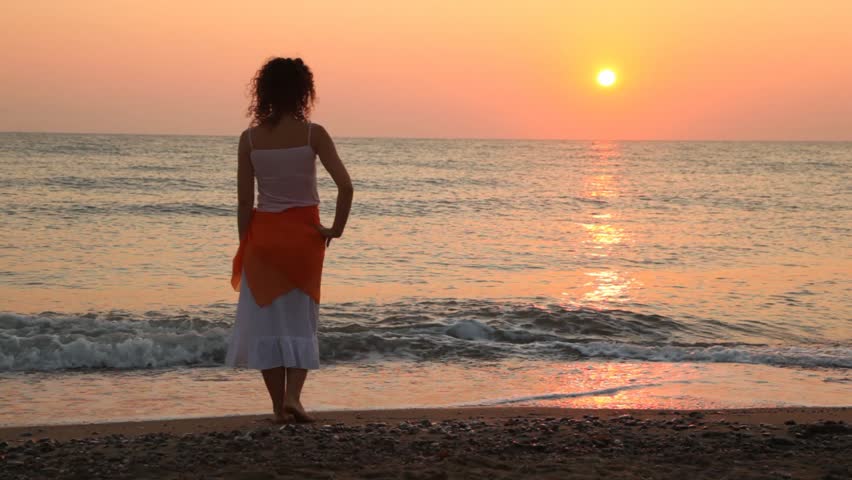 curly-headed woman stands alone on beach faced to sea at sunset