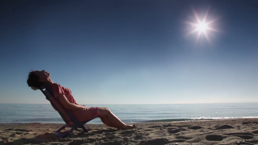 Woman leaned back on deckchair and tans, on beach at sunny day, sideview