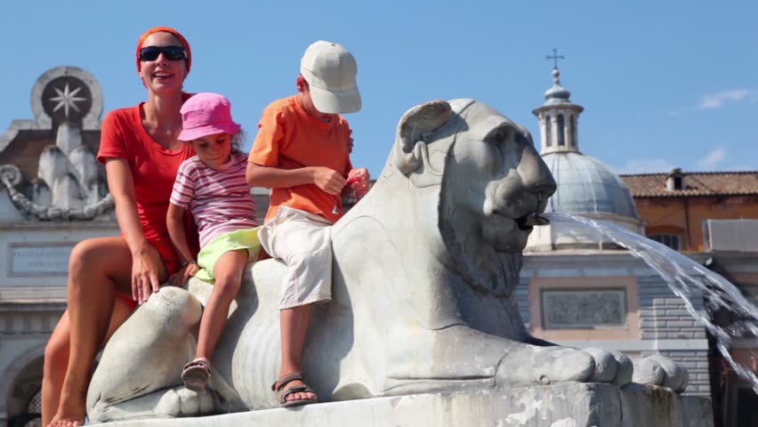 Mother and her kids boy and girl sit on statue of lion near Flaminio obelisk, jet of water pours from the mouth of a lion
