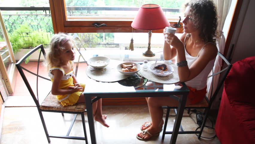 mother and little daughter eat breakfast sitting at table near balcony