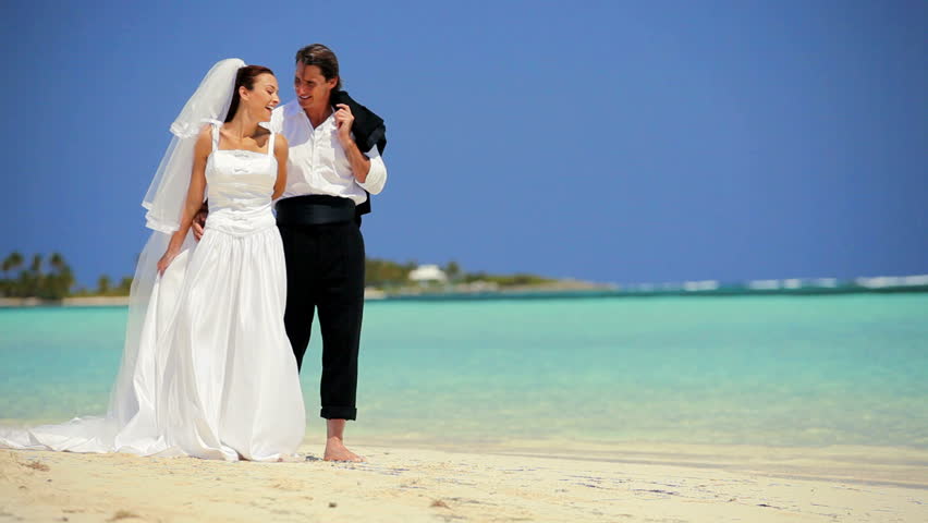 Attractive caucasian couple laughing & dancing barefoot on the beach after their dream wedding