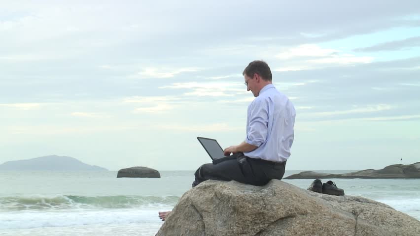 Businessman working with laptop while sitting on a rock on the beach.
