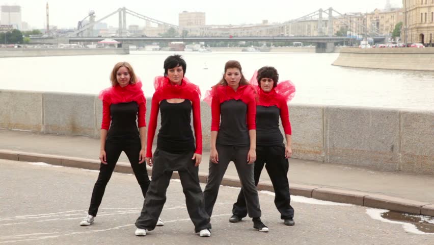 Four girls dance synchronously on quay, then turn around, at background of city traffic on bridge in Moscow