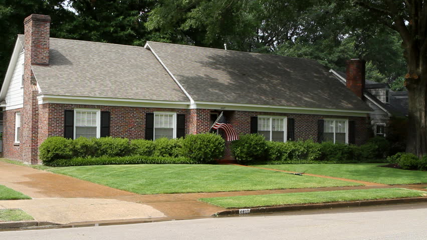 Middle class American brick home in a suburban neighborhood flying an American flag with the lawn sprinkler watering the grass.