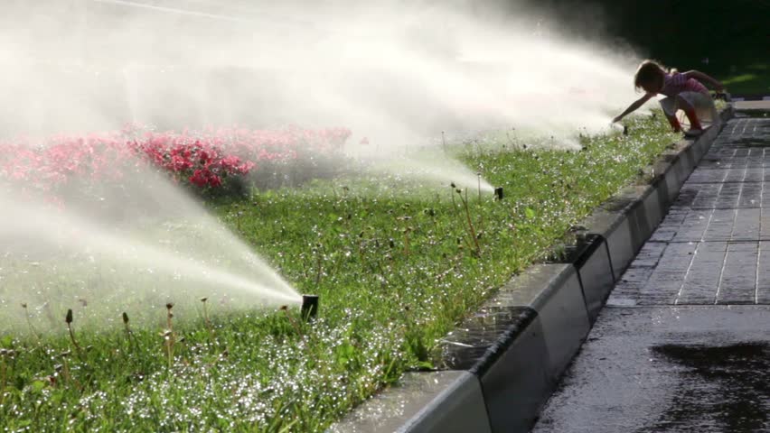 Girl walks along the flowerbed and wets her hands in water, irrigation system abundant watering the plants