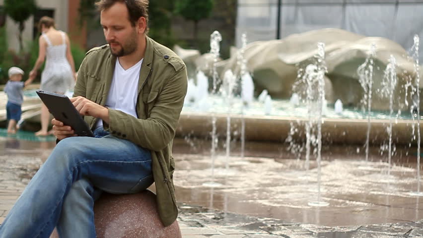 Young happy man with tablet computer sitting by the fountain, slow motion 
