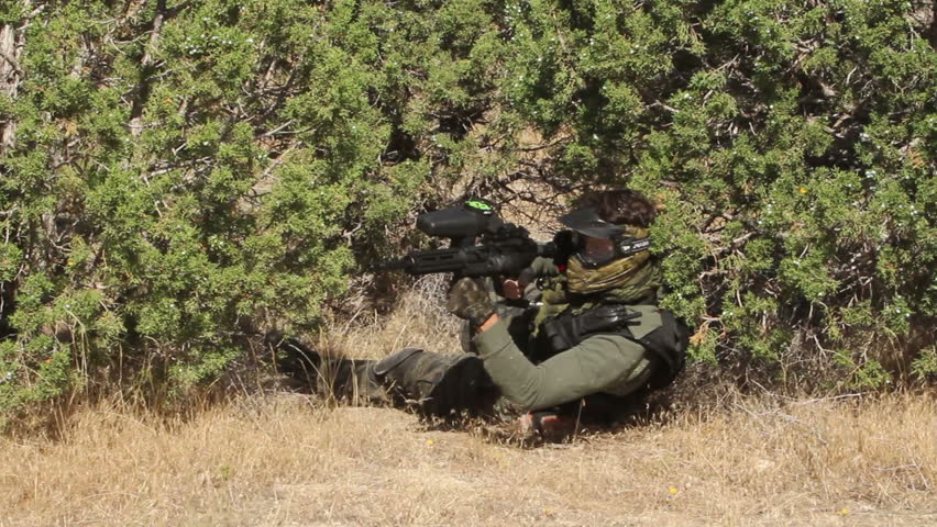 Paintball shooter laying under a tree then running while shooting his fully automatic gun at enemy soldiers. Full camouflage clothes and protective gear.