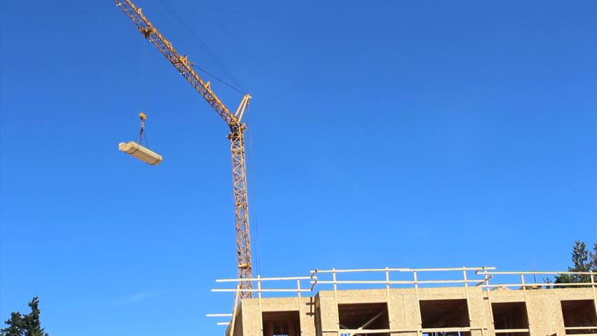 A construction crane pivots carrying a load of lumber at a work site in the suburbs.