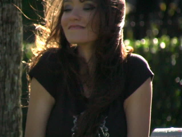 Close-up of a beautiful brunette sitting on a park bench on a breezy summer day. Begins with a close-up of her feet and tilts up.