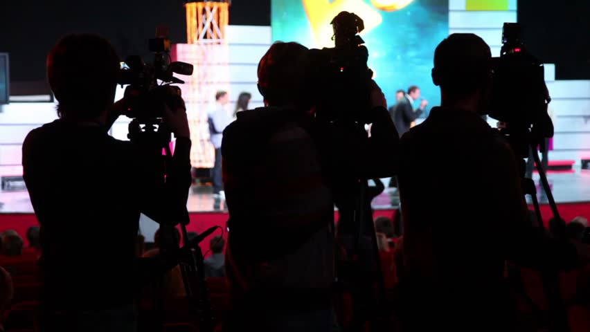 Silhouettes of three operators on workplace in row of few cameras, view from behind on event, people walk on stage
