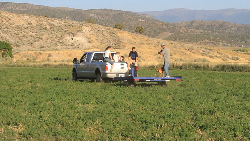 Family and men working on family farm. Men riding trailer to load hay bales. Pickup truck pulling load. Annual agriculture harvest is hard work requiring a lot of heavy lifting.