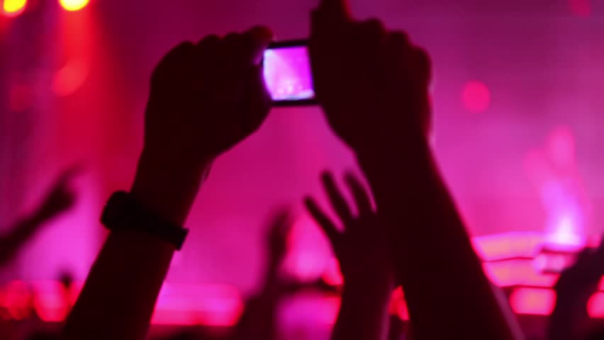 Hands hold camera with digital display among people at rave party with pink red light, view from behind