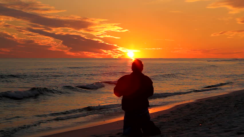 Man kneeling at the beach as the sun sets on the horizon and raises his arms in worship.