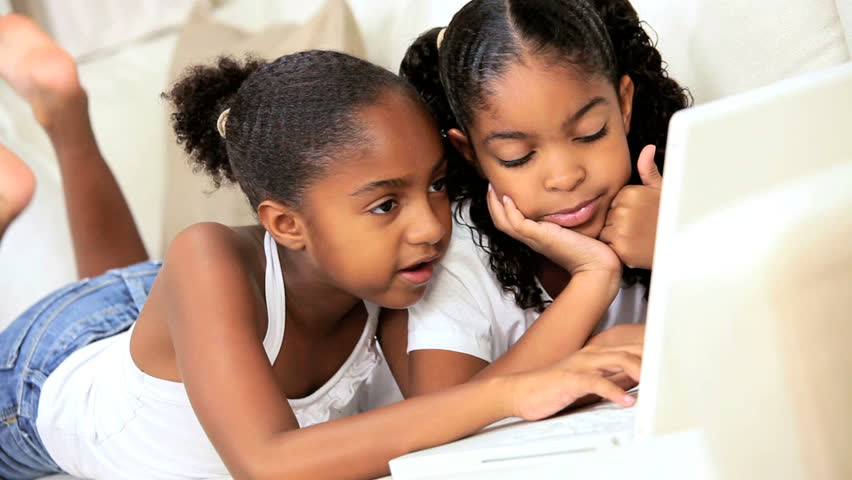 Cute little african american girls lying on their home sofa playing internet games on a laptop