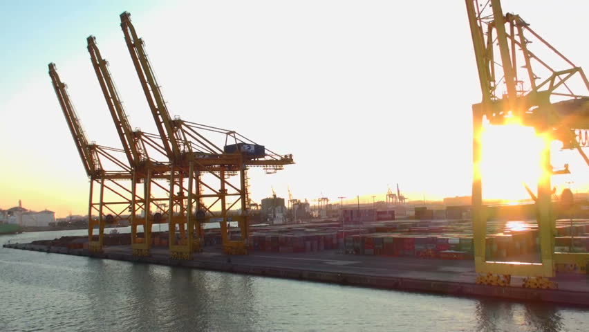 Ship leaving Barcelona harbor sailing along the jetty pier with its many crane spreaders and containers, sunset time.