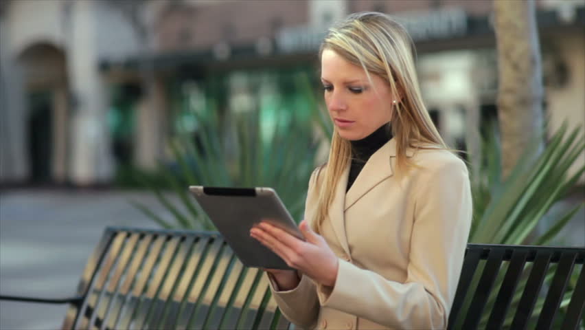 Sitting on a municipal park bench using an electronic tablet a pretty blond businesswoman enjoys the sunshine of a breezy fall day.
