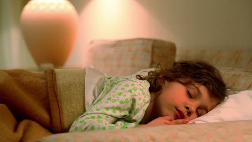 Little girl lies in bed and rubs her eyes at background of lamp