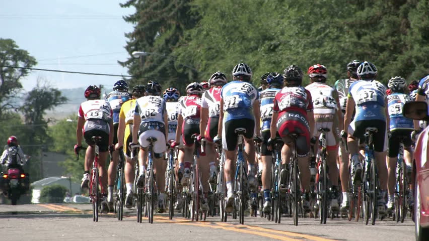 Tour of Utah bicycle race. Group riding away. Coming into a small town of Chester in Sanpete County. Group of riders at high speed. Motorcycle with judges and support cars following. 