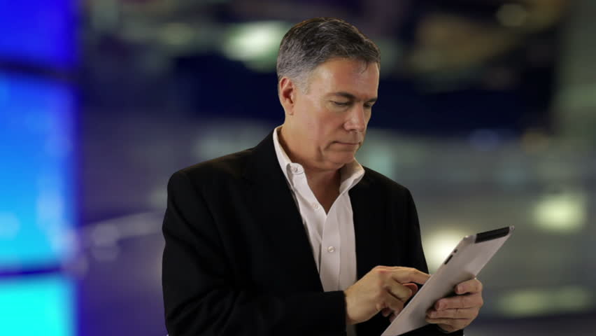 A handsome mature man standing in a large room with strange lights stops looking at his digital tablet and smiles.