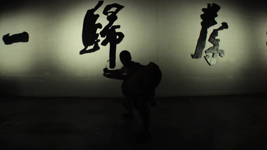 A Shaolin monk demonstrates in silhouette an ancient form of kung fu using a practice sword and shield. Shot in slow motion. 