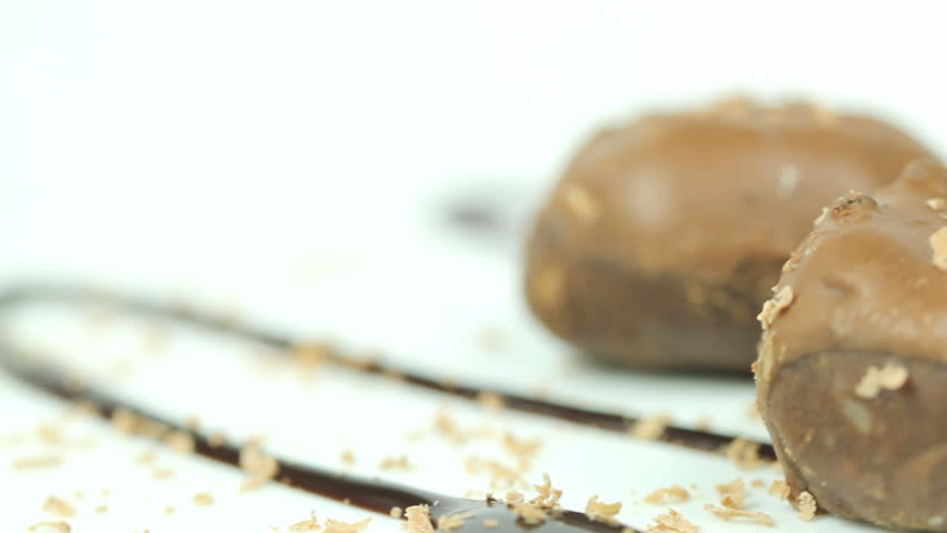 Two chocolate covered coffee flavoured cookies with hazelnut chips rotating on a white plate covered in chocolate syrup and bits of light milk chocolate