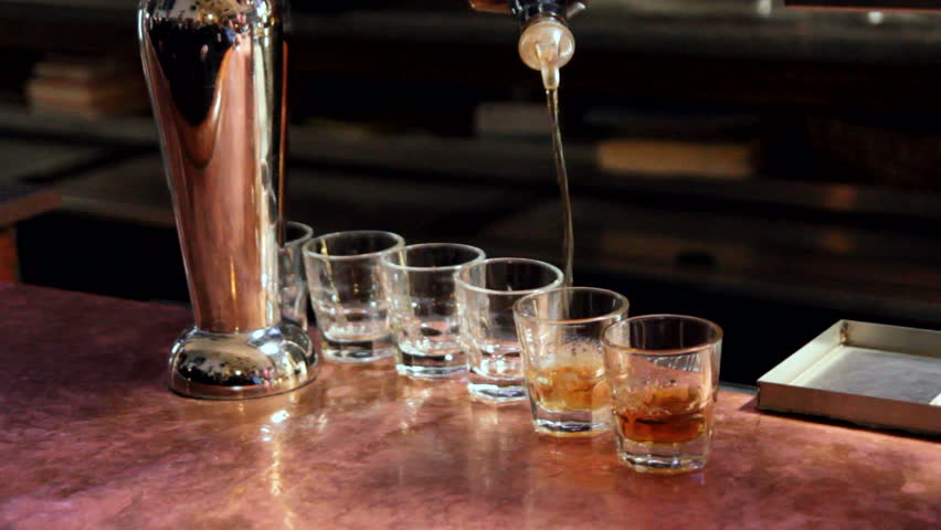 Bar tender pouring whiskey into six shot glasses.