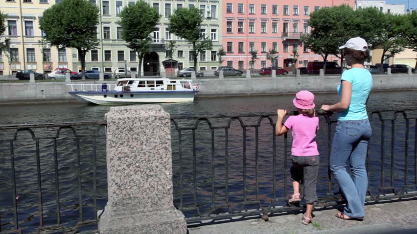 Woman in white cap and sunglasses stand with her daughter on quay near fence, city traffic is at other side of Fontanka river