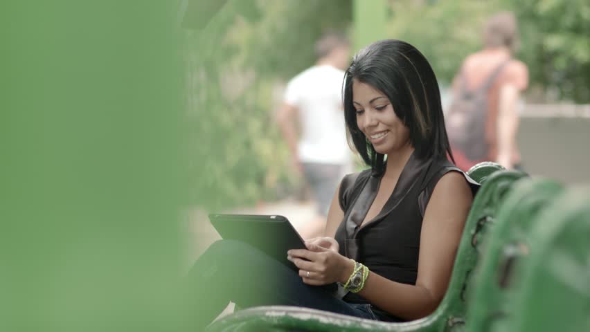 Portrait of happy young hispanic woman typing on digital tablet computer and sitting on bench in city park