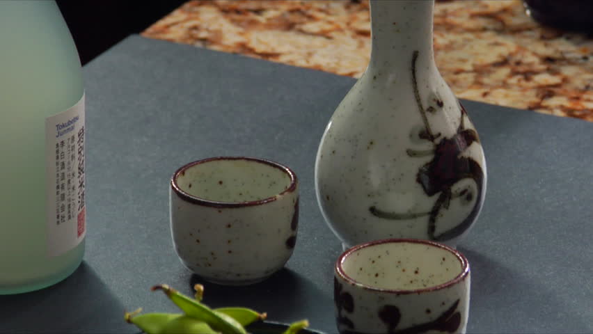 Close-up Pan down of steaming hot cups of sake and man sprinkling salt on Edamame (soy beans).