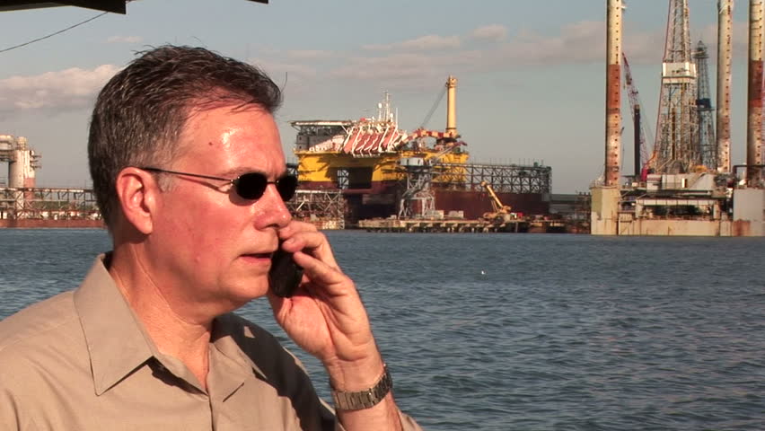 A close up of a man on a cell phone with oilrigs and other offshore platforms in the background