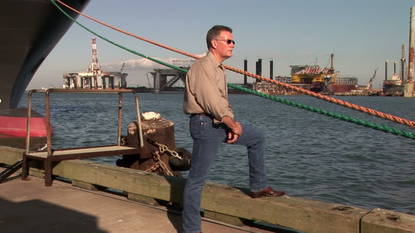 A man standing on a pier with oilrig and other offshore platforms in the background answers a call.