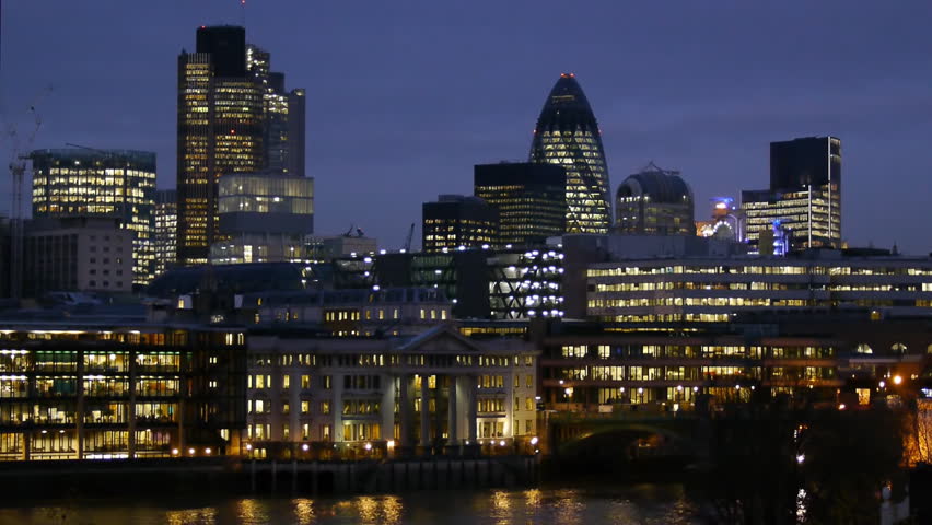 Skyline of the office towers of the City of London at night. Buildings include the Gerkin, Nat West Tower, Lloyds of London. The river Thames is visible at the bottom of the frame.