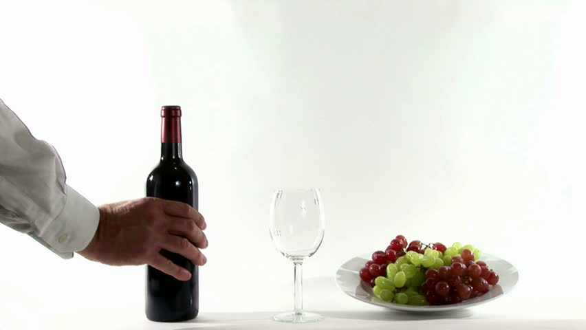 A man pours a glass of red wine with a plate of red and white grapes accenting the scene.