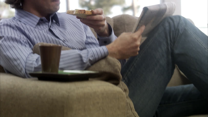 Man sitting on sofa reading newspaper and having breakfast