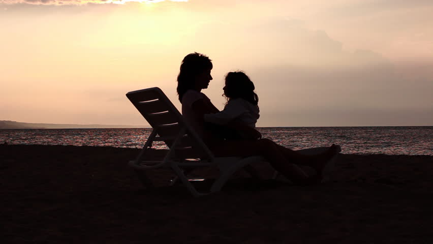 Mother and daughter hugging and kissing on a lounge by the sea. Sunrise in the background
