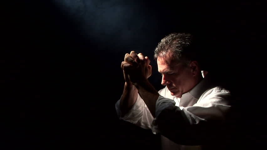 A man spot lighted by a fog filled beam of light, kneeling in a posture of prayer or supplication.