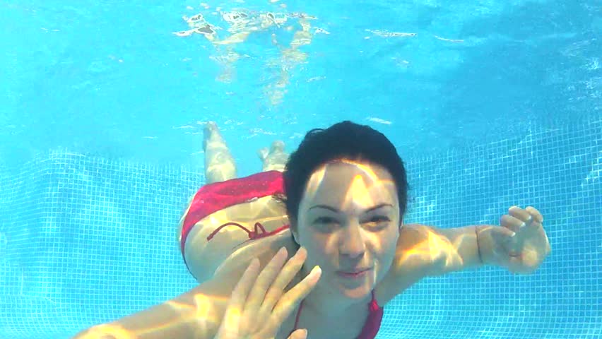 Underwater view as woman swims towards camera and waves as she blows bubbles.