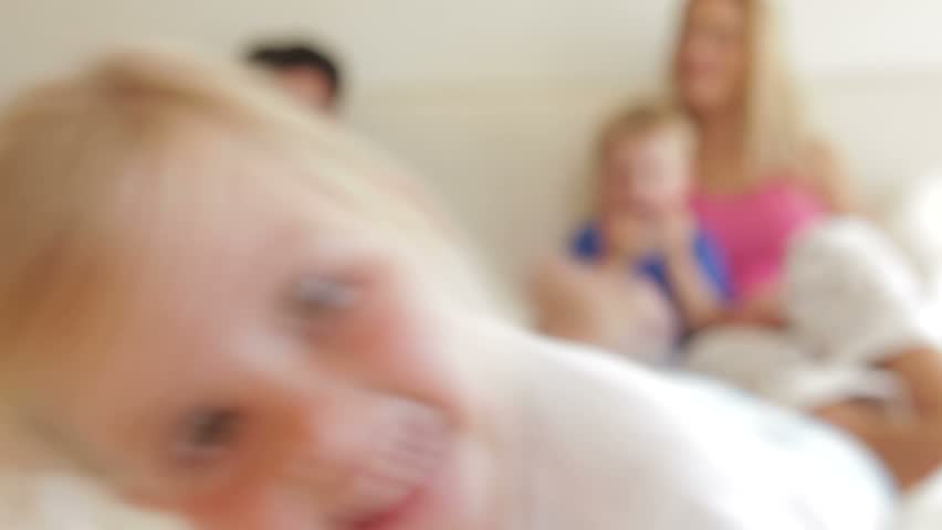 Shallow focus view of family sitting in bed, daughter crawls towards camera and looks into the lens.