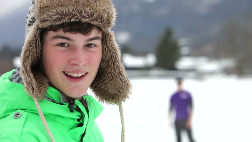 Teenage boy faces camera, tosses snowball in the air and then throws it at his friend in the distance.