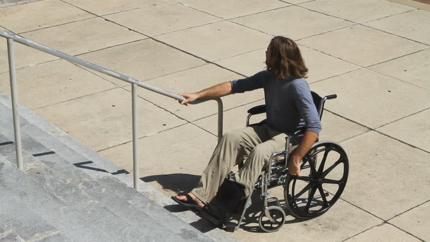Man in a wheelchair rolls up to a concrete stairway looking for a ramp to access the building and throws up his arms in frustration.
