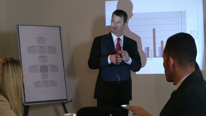 Action tracks from a businessman standing in front of an image projected onto him by a projector to a man in the room asking a question. 
