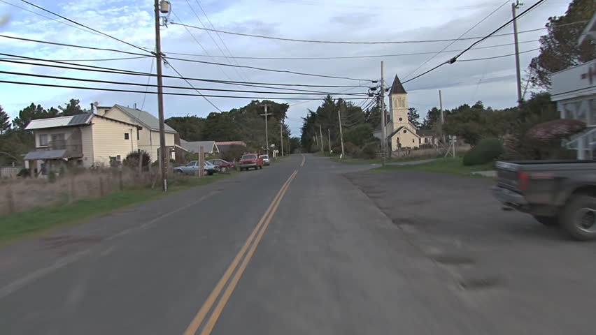 First person driving POV shot moving north through the town of Caspar, California, under power lines toward an old church with classic steeple.
