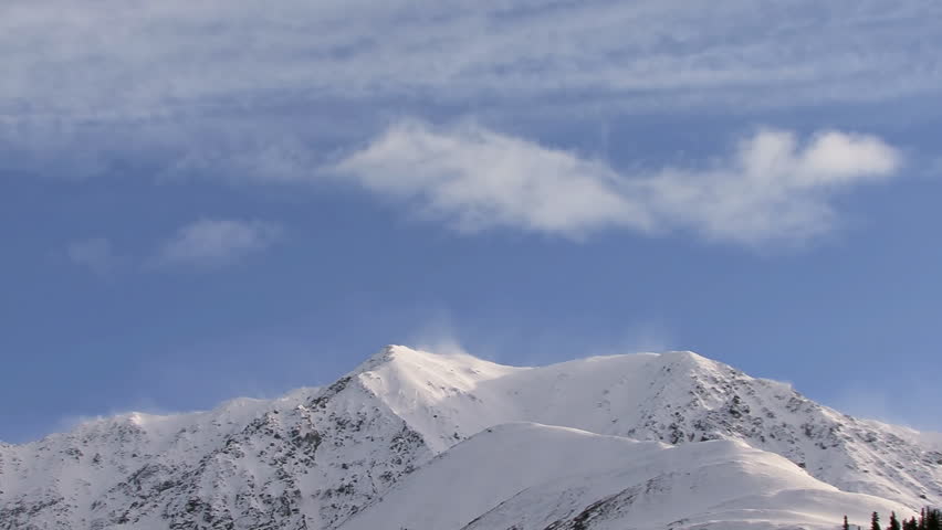 Stark, craggy mountains, with the air currents slamming into the other side forcing spindrift and billowing clouds to forma and dissipate over the mountain's edge. Time lapse.